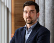 Ein Mann mit kurzen dunklen Haaren und Bart, der ein blaues Hemd und einen dunklen Blazer trägt, steht vor einem Fenster. Vor dem Hintergrund der hölzernen und grauen Wände der Hochschule Coburg behält er einen neutralen Gesichtsausdruck bei.