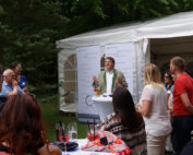 Eine Gruppe von Menschen versammelt sich im Freien in der Nähe eines weißen Zeltes und hört einem Vortragenden an einem kleinen runden Tisch zu. Im Hintergrund sind Banner zu sehen, darunter eines für die Hochschule Coburg. Auf den Tischen stehen verschiedene Getränke, Bäume umgeben den Bereich.