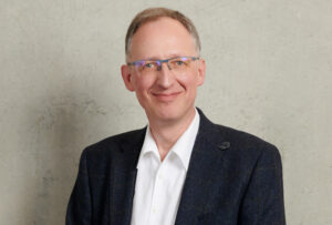 A smiling man with short hair and glasses, donning a dark blazer and white shirt, stands against a light textured background, embodying the professionalism associated with Hochschule Coburg.