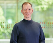 Eine Person mit kurzen Haaren steht lächelnd im Innenbereich der Hochschule Coburg. Sie trägt ein dunkles Langarmshirt und im Hintergrund ist ein großes Fenster mit Blick auf das üppige grüne Laub draußen zu sehen.