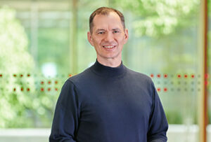 A person in a dark long-sleeve shirt stands indoors at Hochschule Coburg, smiling at the camera. The blurred green background includes a glass window adorned with a line of small decorative dots.