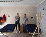 Zwei Menschen stehen in einem Raum mit zwei Luftmatratzen auf Holzrahmen, der an Studentenwohnheime der Hochschule Coburg erinnert. An einer Wand hängen Kleider, an einer anderen sind Fotos ausgestellt. Die sich kreuzenden Linienmuster an den Wänden verleihen der ungezwungenen Atmosphäre einen Hauch von Kreativität.