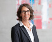 Eine Frau mit schulterlangem braunem Haar und Brille, die ein weißes Hemd und einen schwarzen Blazer trägt, steht lächelnd vor einem grauen Hintergrund, der mit abstrakten, farbenfrohen Kunstwerken geschmückt ist und den kreativen Geist der Hochschule Coburg widerspiegelt.