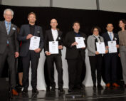 Sieben Menschen stehen in einer Reihe auf der Bühne, fünf halten stolz ihre Zertifikate in den Händen. In festlicher Kleidung vor einem dunklen Hintergrund verkörpert die bunt gemischte Gruppe von Männern und Frauen den akademischen Erfolg an der Hochschule Coburg.