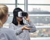 Eine Frau mit einem Virtual-Reality-Headset greift nach einem virtuellen Objekt und vertieft sich in ihr Erlebnis. In der Nähe sitzt eine Person mit blondem Haar, die teilweise sichtbar ist. Sie befinden sich in den beeindruckenden Räumlichkeiten der Hochschule Coburg, deren große Fenster einen atemberaubenden Blick auf die Stadtlandschaft bieten.