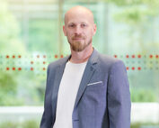 Ein kahlköpfiger Mann mit Bart steht selbstbewusst in einem Innenraum der Hochschule Coburg. Er trägt ein weißes Hemd und einen grauen Blazer, während große Fenster hinter ihm den Blick auf das üppige Grün draußen freigeben.