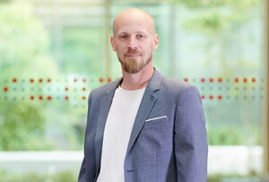 A man with a bald head and beard, wearing a gray blazer and white shirt, stands in front of a softly focused background with greenery at Hochschule Coburg. His expression is neutral, and he is positioned slightly to the left.