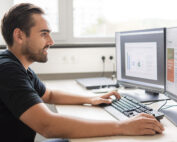 Ein Mann sitzt an einem Schreibtisch und konzentriert sich auf zwei Computermonitore, auf denen Grafiken und Texte angezeigt werden. Durch das Fenster hinter ihm fällt Tageslicht in seinen Arbeitsbereich. Als Absolvent der Hochschule Coburg wendet er sein erlerntes Fachwissen an, um Daten mithilfe von Tastatur und Maus effizient zu analysieren.
