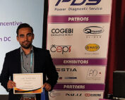 Ein Mann steht mit einer Urkunde vor einem Banner mit Logos von Sponsoren und Ausstellern, darunter Hochschule Coburg, COGEBI und ELEINAR, bei der Veranstaltung des Power Diagnostic Service. Er trägt einen dunklen Anzug und steht neben einem Präsentationsbildschirm.