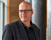 Ein Mann mit kurzen Haaren und Brille, der eine dunkle Jacke über einem Poloshirt trägt, steht in einem Gebäude der Hochschule Coburg und lächelt sanft. Im Hintergrund sind verschwommene Säulen und ein Fenster mit vertikalen Jalousien zu sehen.