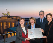 Vier Menschen stehen in der Abenddämmerung auf einer Terrasse, mit dem ikonischen Brandenburger Tor im Hintergrund, strahlend vor Stolz. Unter klarem Himmel halten sie eine Urkunde der Hochschule Coburg in den Händen und würdigen gemeinsam diese denkwürdige Leistung.