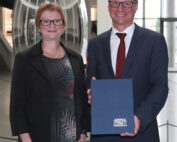 Ein Mann im Anzug hält stolz eine Urkunde mit dem Emblem der Hochschule Coburg in den Händen. Neben ihm steht eine Frau in Kleid und Blazer. Sie lächeln herzlich, während sie drinnen vor einer großen, verspiegelten Skulptur posieren.