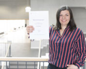 Eine Person in einem gestreiften Hemd steht in einem Innenbereich der Hochschule Coburg, lächelt und hält mit beiden Händen ein Dokument hoch. Die Umgebung wirkt modern mit natürlichem Licht und minimalistischem Design.