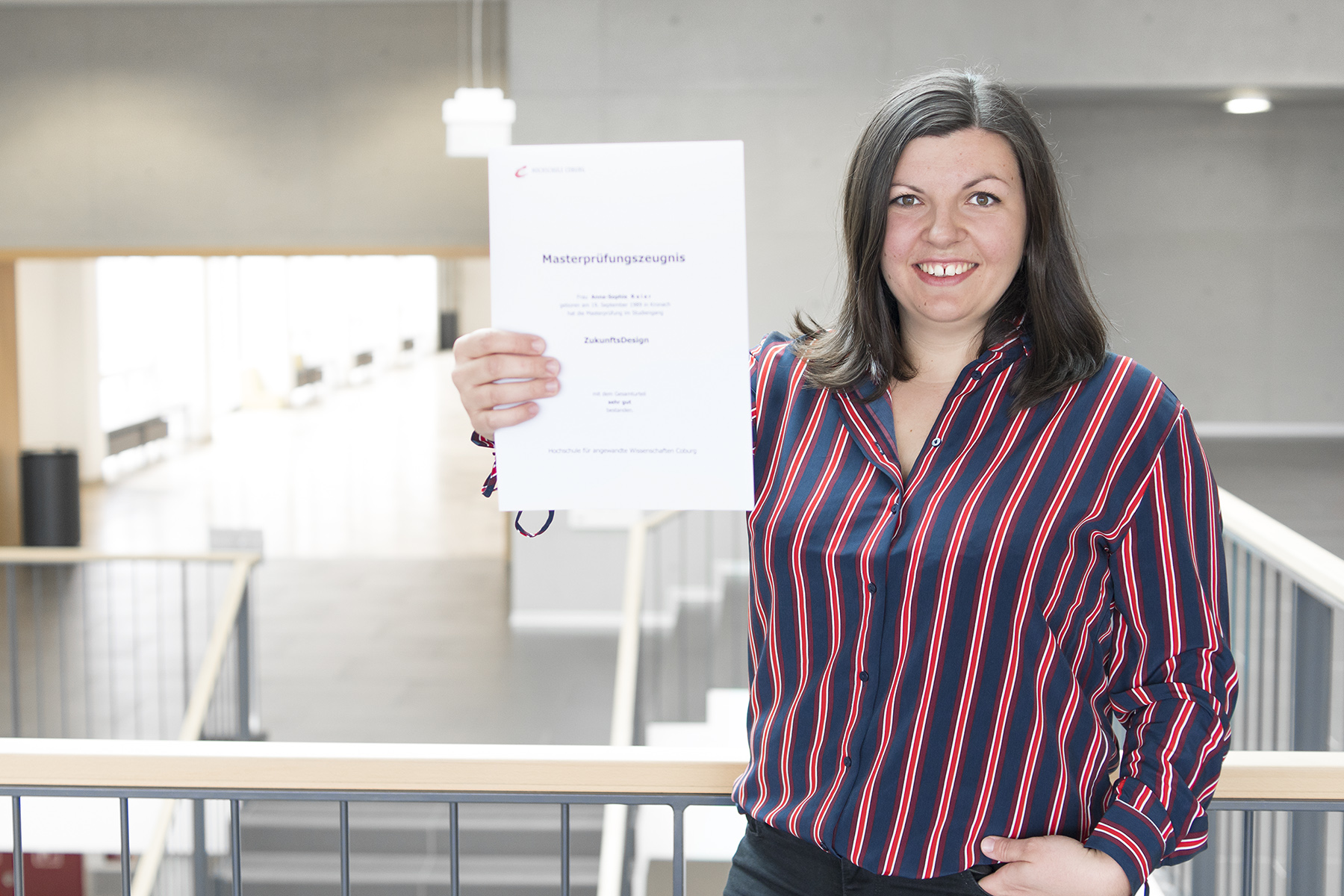 Eine Person in einem gestreiften Hemd steht in einem Innenbereich der Hochschule Coburg, lächelt und hält mit beiden Händen ein Dokument hoch. Die Umgebung wirkt modern mit natürlichem Licht und minimalistischem Design.