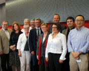 Eine Gruppe von zwölf Personen, sowohl Männer als auch Frauen, posiert für ein Foto im Innenbereich der Hochschule Coburg. Sie sind in formeller und legerer Business-Kleidung gekleidet, im Hintergrund sind eine gestreifte Wand und ein rotes Paneel zu sehen.