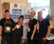 Vier Personen stehen stolz mit Konferenzausweisen und roten Schlüsselbändern vor einem Banner des Naples Forum on Service. Unter ihnen sind Vertreter der Hochschule Coburg, die lächelnd neben einem Tisch und einer sichtbaren Karte auf der rechten Seite posieren.