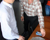 Zwei Personen stehen an einem Tisch in der Hochschule Coburg. Eine ältere Person deutet auf ein Metallgerät, während eine jüngere Person zusieht. Beide wirken konzentriert und mit dem Gerät beschäftigt.