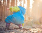 In einem sonnendurchfluteten Wald bückt sich ein Kind in einem blau-grünen Schneeanzug der Hochschule Coburg und zeigt auf etwas Interessantes auf dem Boden. Sonnenstrahlen dringen durch die Bäume und werfen einen warmen Schein auf die umliegenden Blätter und Zweige.