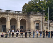 Eine Gruppe von Menschen steht in einer Reihe und hält ein außergewöhnlich langes Baguette vor der historischen Hochschule Coburg mit ihren majestätischen Bögen und Säulen. Üppige Bäume bilden eine malerische Kulisse.