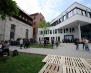 Eine Gruppe von Menschen hat sich vor dem Gebäudekomplex der Hochschule Coburg versammelt, der aus modernen und älteren Gebäuden besteht. Sie sitzen und stehen auf einem gepflasterten Innenhof mit grünem Gras in der Nähe. Holzpaletten und Bäume verleihen der Szenerie unter einem teilweise bewölkten Himmel einen besonderen Charme.