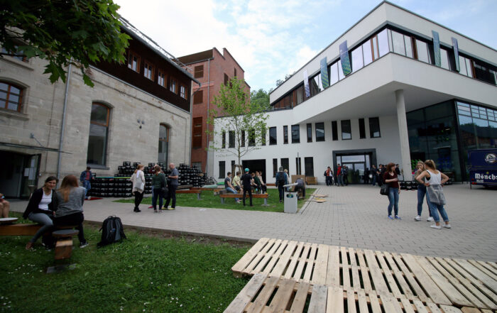 Eine Gruppe von Menschen hat sich vor dem Gebäudekomplex der Hochschule Coburg versammelt, der aus modernen und älteren Gebäuden besteht. Sie sitzen und stehen auf einem gepflasterten Innenhof mit grünem Gras in der Nähe. Holzpaletten und Bäume verleihen der Szenerie unter einem teilweise bewölkten Himmel einen besonderen Charme.
