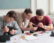 Drei junge Männer arbeiten gemeinsam an einem Tisch in der Hochschule Coburg an einem Elektronikprojekt. In einem hellen Raum mit großen Fenstern löten sie konzentriert Bauteile, die mit Drähten verbunden sind, umgeben von Werkzeug und Materialien.