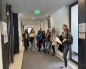 Eine Gruppe von Menschen steht in einem Flur der Hochschule Coburg, einige lächeln und halten Papiere in den Händen. Über ihnen leuchten helle Lichter und Notausgangsschilder. Die Menge wirkt engagiert und aufmerksam, es handelt sich um eine bunte Mischung aus Männern und Frauen in legerer Kleidung.