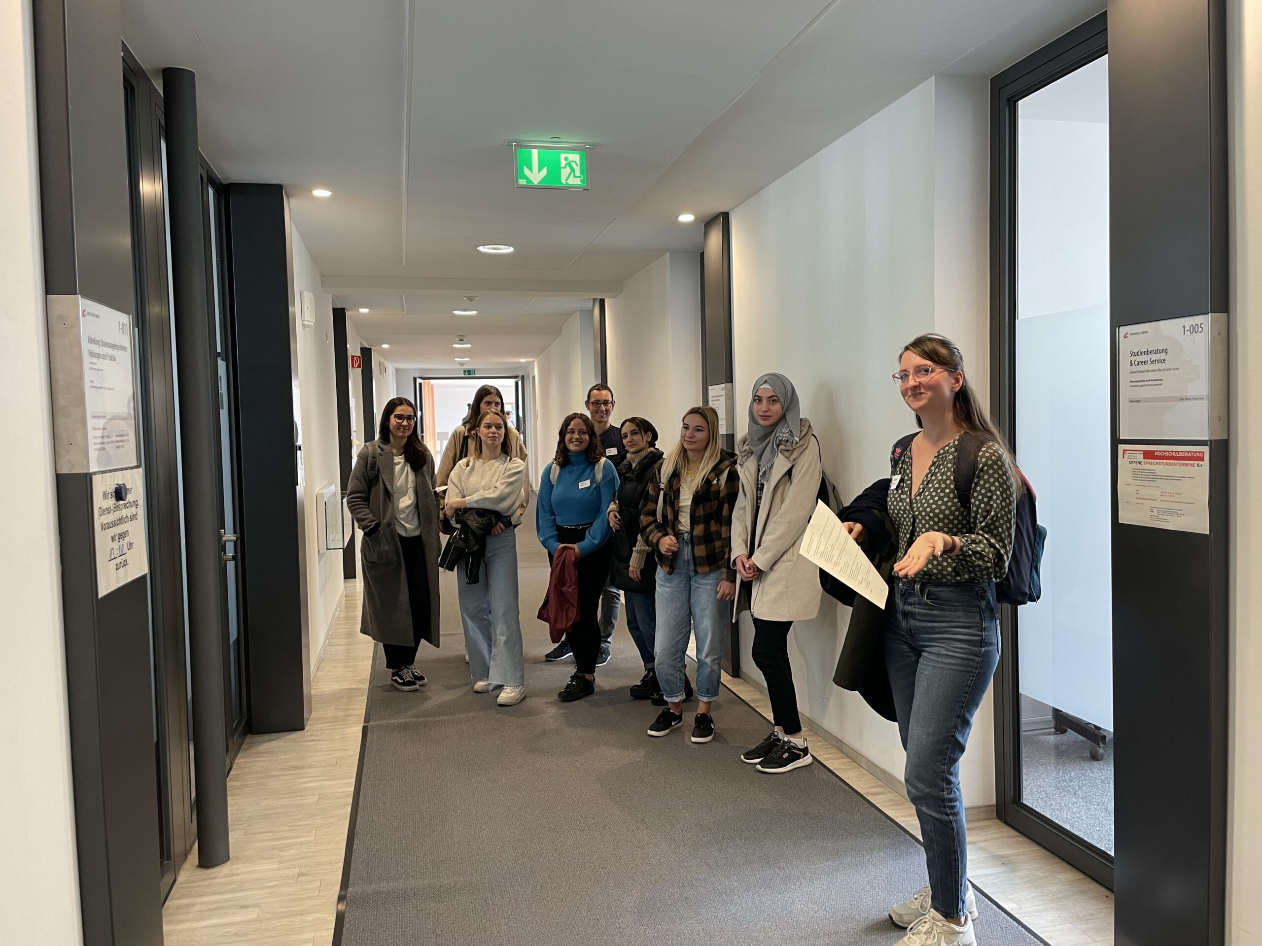 Eine Gruppe von Menschen steht in einem Flur der Hochschule Coburg, einige lächeln und halten Papiere in den Händen. Über ihnen leuchten helle Lichter und Notausgangsschilder. Die Menge wirkt engagiert und aufmerksam, es handelt sich um eine bunte Mischung aus Männern und Frauen in legerer Kleidung.