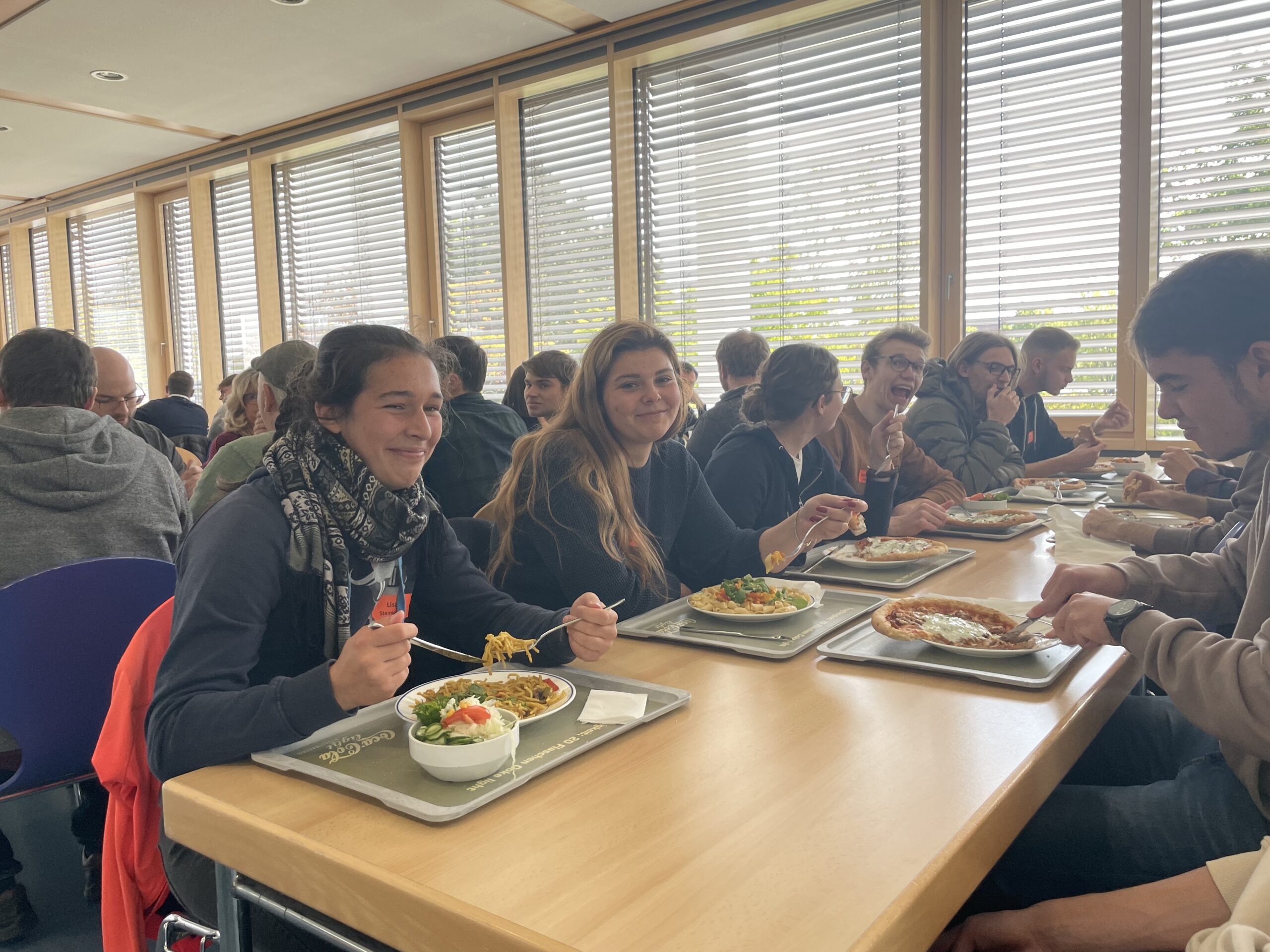 Eine Gruppe von Menschen sitzt an einem langen Holztisch in der Cafeteria der Hochschule Coburg und genießt Mahlzeiten auf Tabletts. Durch die großen Fenster mit Jalousien fällt natürliches Licht in den Raum und schafft eine warme Atmosphäre. Alle scheinen in ein Gespräch und Essen vertieft zu sein.