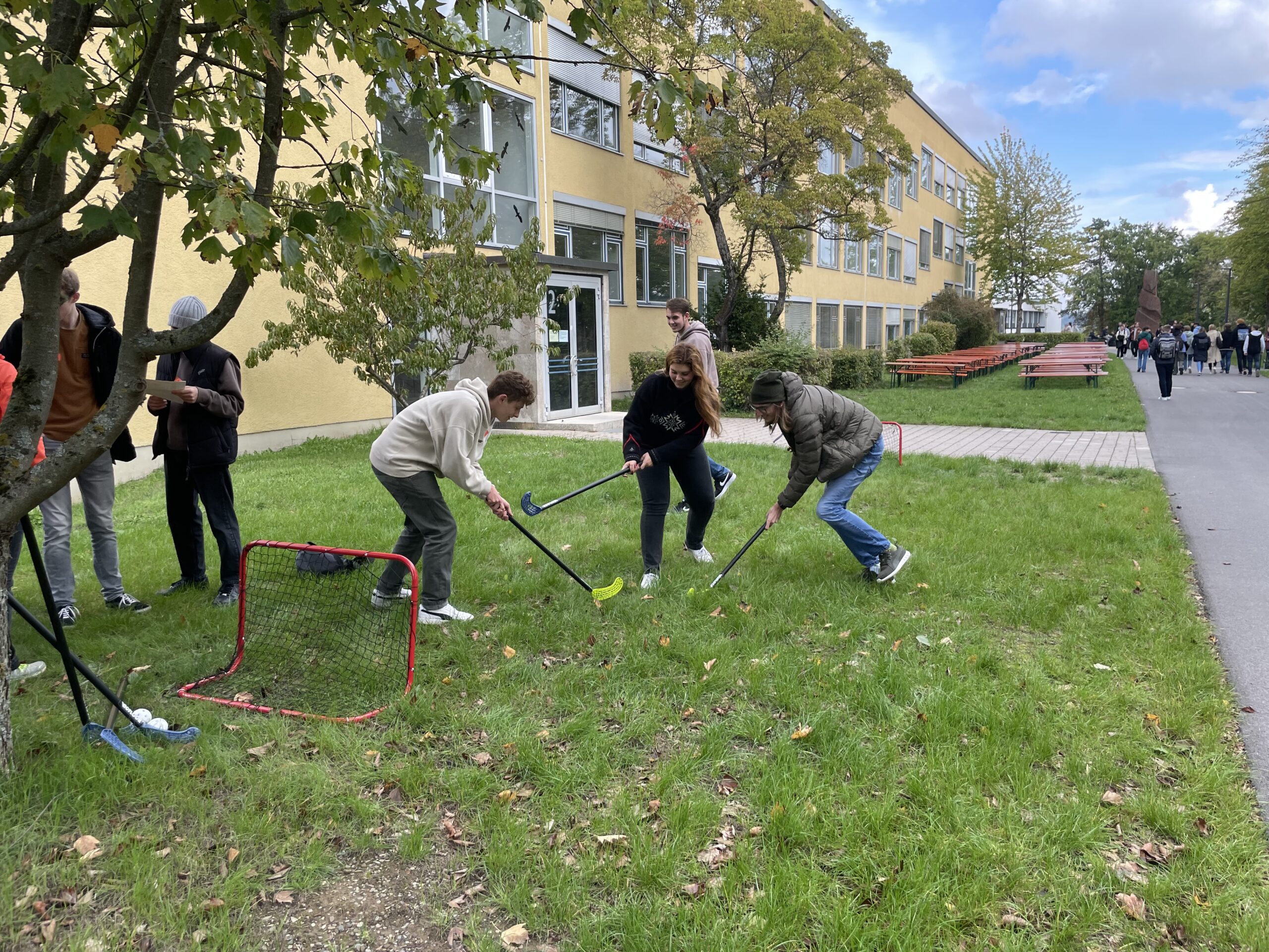 Eine Gruppe von Leuten der Hochschule Coburg spielt Unihockey auf einer Rasenfläche neben einem Gebäude. Sie versammeln sich um ein kleines Netz, halten Schläger in der Hand und spielen ein freundschaftliches Spiel. Bäume und ein Weg vervollständigen die malerische Kulisse.