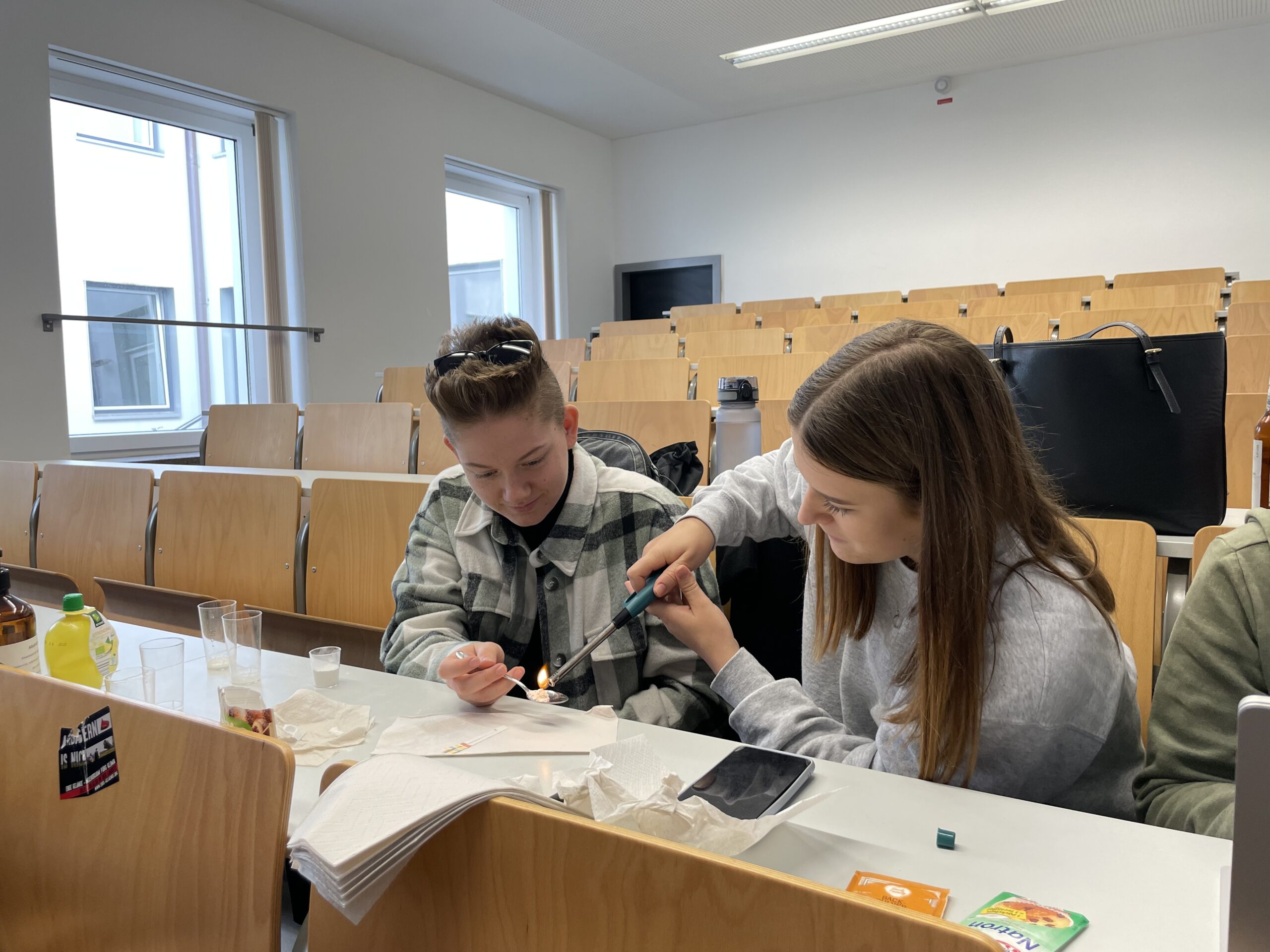Zwei Personen arbeiten in einem Hörsaal der Hochschule Coburg gemeinsam an einer Aktivität. Einer benutzt ein Werkzeug an einem kleinen Gegenstand, den der andere hält, umgeben von Papieren, Getränken und verschiedenen Gegenständen auf dem Schreibtisch. Der Raum verfügt über Holzsitze und große Fenster, durch die natürliches Licht hereinströmt.
