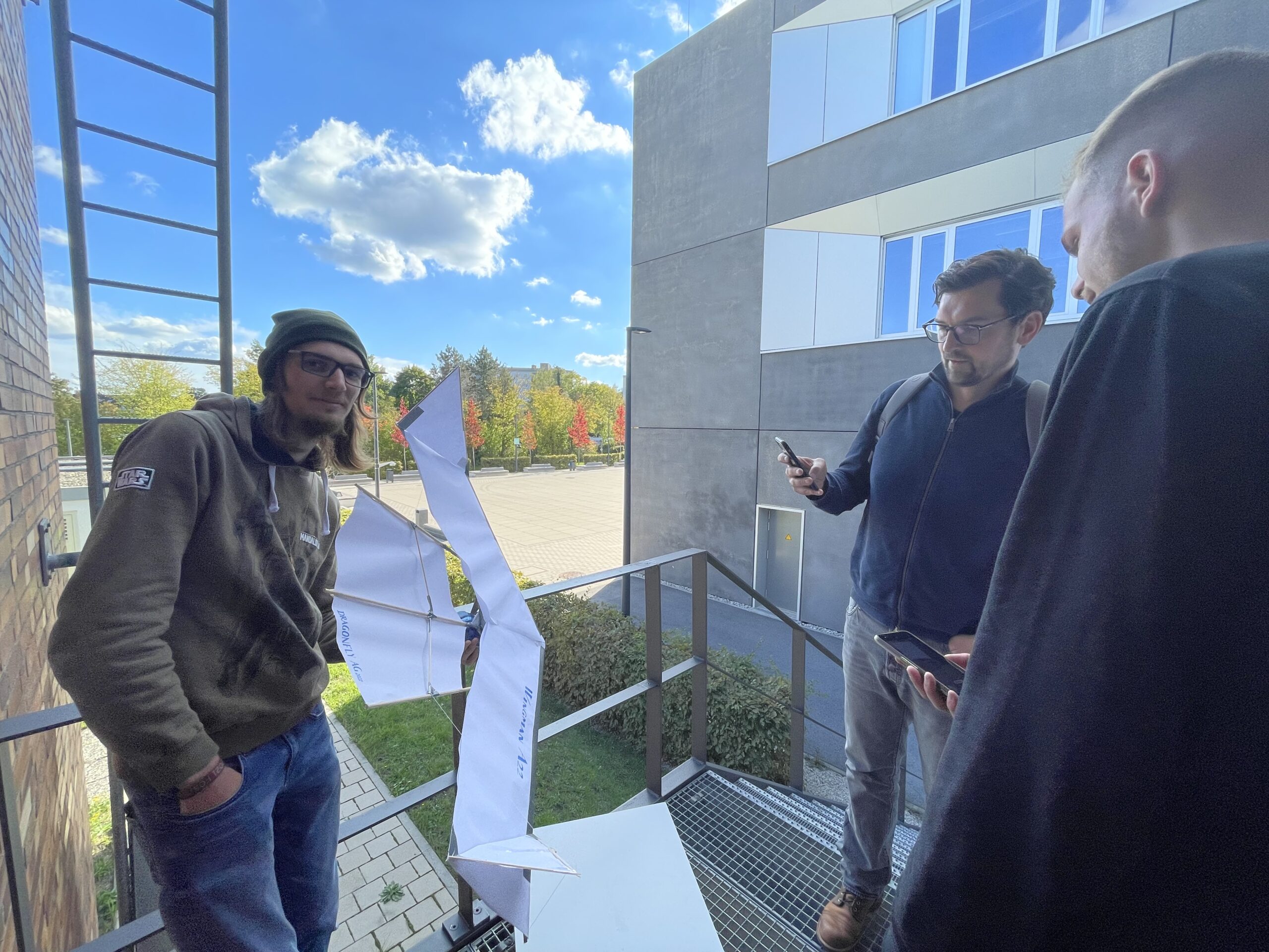 Drei Personen auf einem Balkon der Hochschule Coburg untersuchen ein großes Papiermodell einer weißen Struktur. Eine Person mit Hut steht links, während die anderen beiden Geräte halten. Ein sonniger Tag zeigt einen Parkplatz und im Hintergrund herbstliche Bäume.