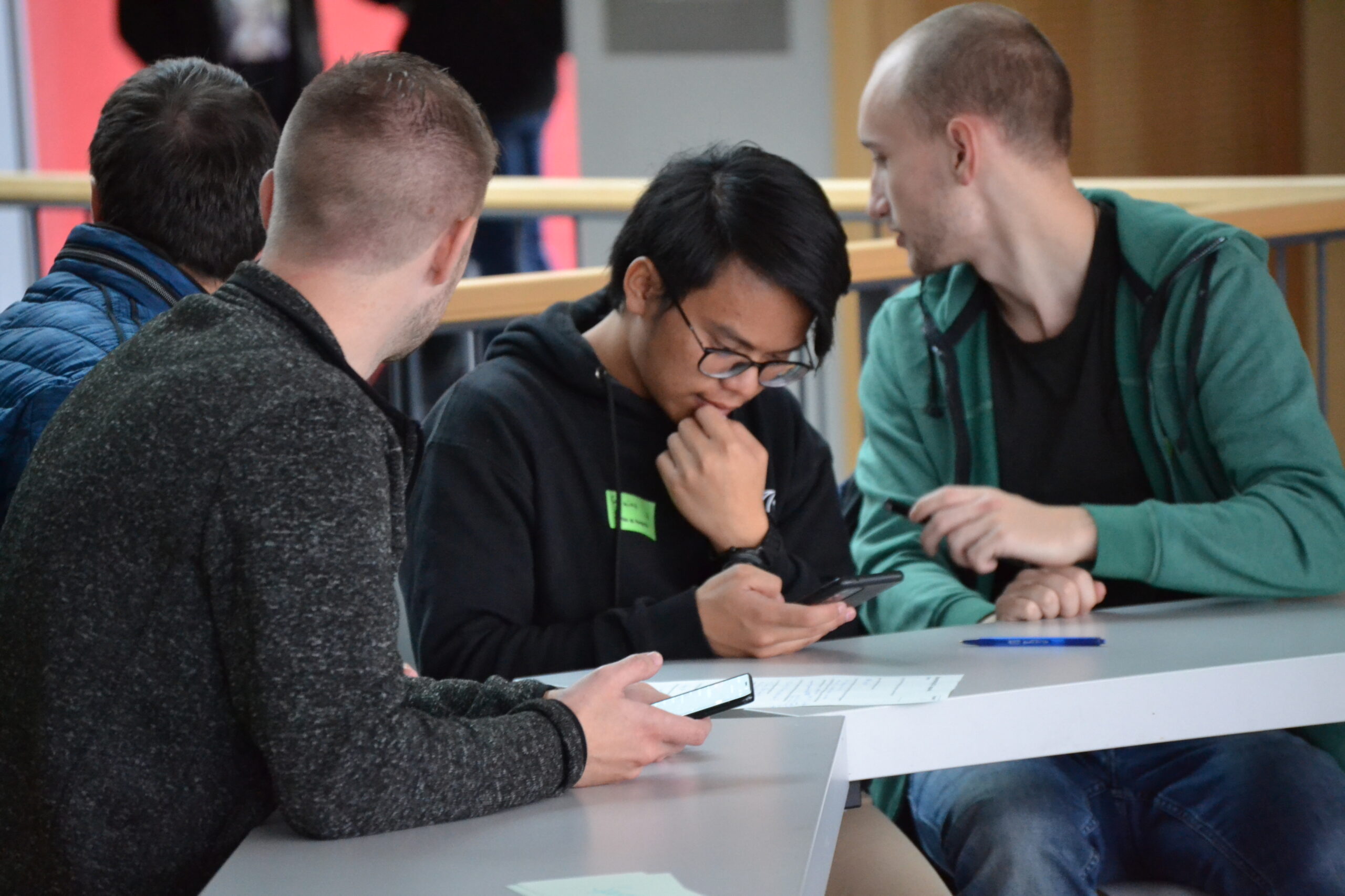 Drei Personen sitzen an einem Tisch in der Hochschule Coburg, zwei konzentrieren sich auf ihre Telefone, während einer einen Aufsatz liest. Sie sind leger gekleidet und unterhalten sich oder lernen. Ein Geländer und ein unscharfer Hintergrund lassen auf die geschäftige Atmosphäre dieser lebendigen akademischen Umgebung schließen.