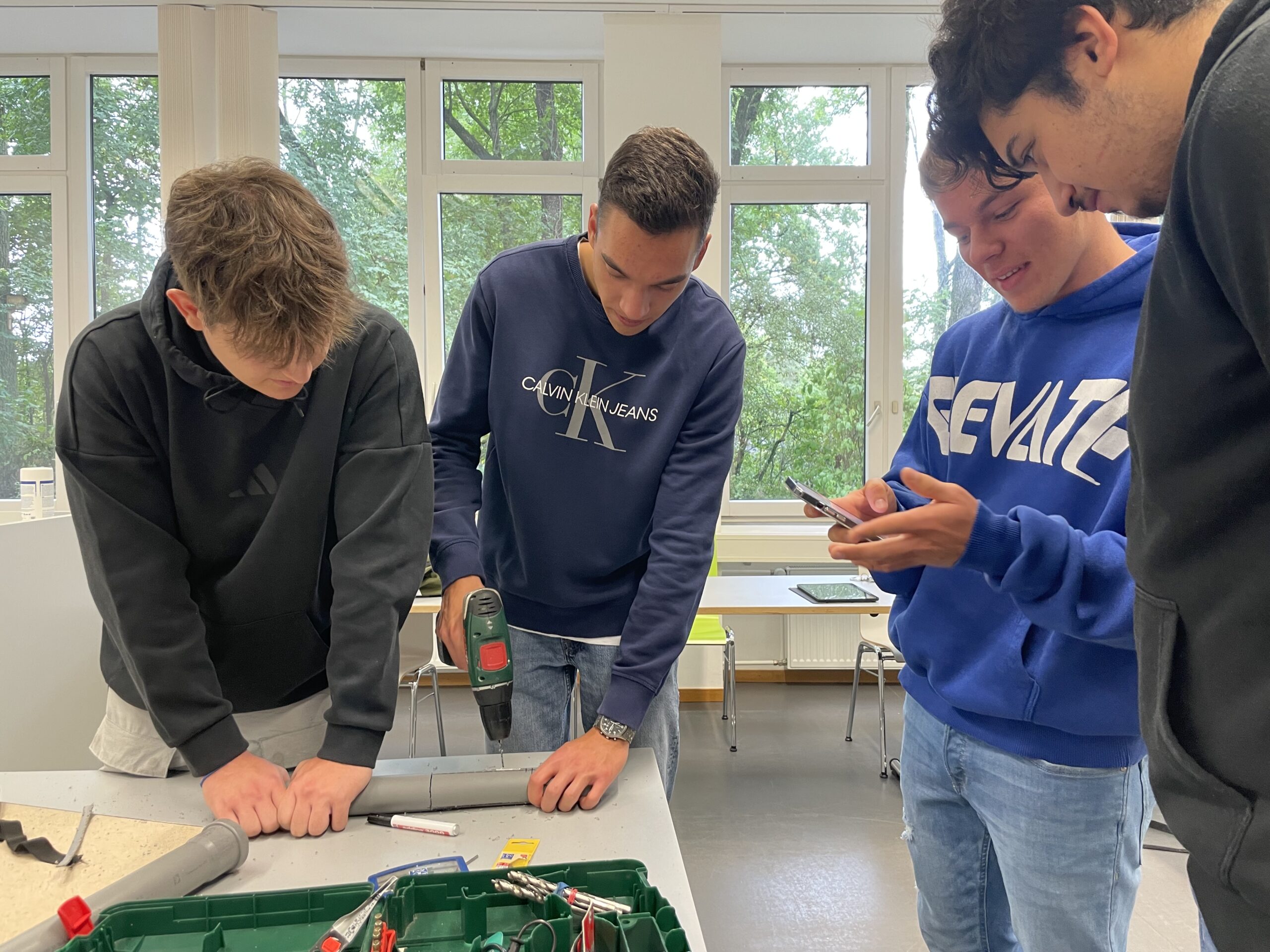 Vier junge Männer der Hochschule Coburg sitzen um einen Tisch. Einer bohrt geschickt mit einer elektrischen Bohrmaschine ein graues Rohr, die anderen schauen aufmerksam zu, einer hält ein Smartphone in der Hand. Sie arbeiten in einem hellen Raum mit großen Fenstern, die die üppigen grünen Bäume draußen einrahmen.