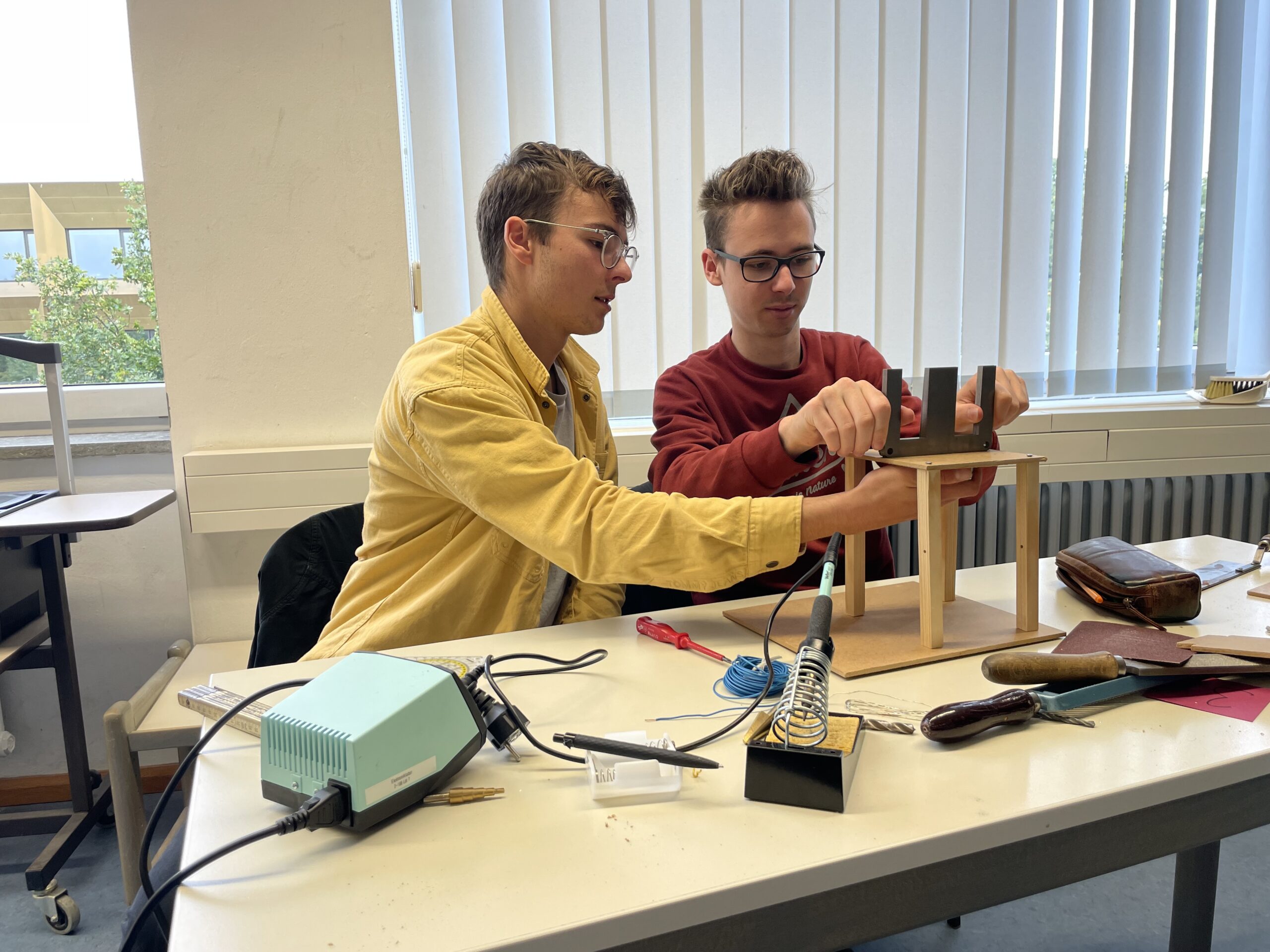 Zwei junge Männer sitzen an einem Tisch mit technischen Geräten und arbeiten konzentriert an einem kleinen Projekt. Der Arbeitsplatz an der Hochschule Coburg ist vollgestopft mit Werkzeugen, Notizbüchern und einer Lötstation. Sie sind konzentriert dabei, eine Holzkonstruktion zusammenzusetzen.