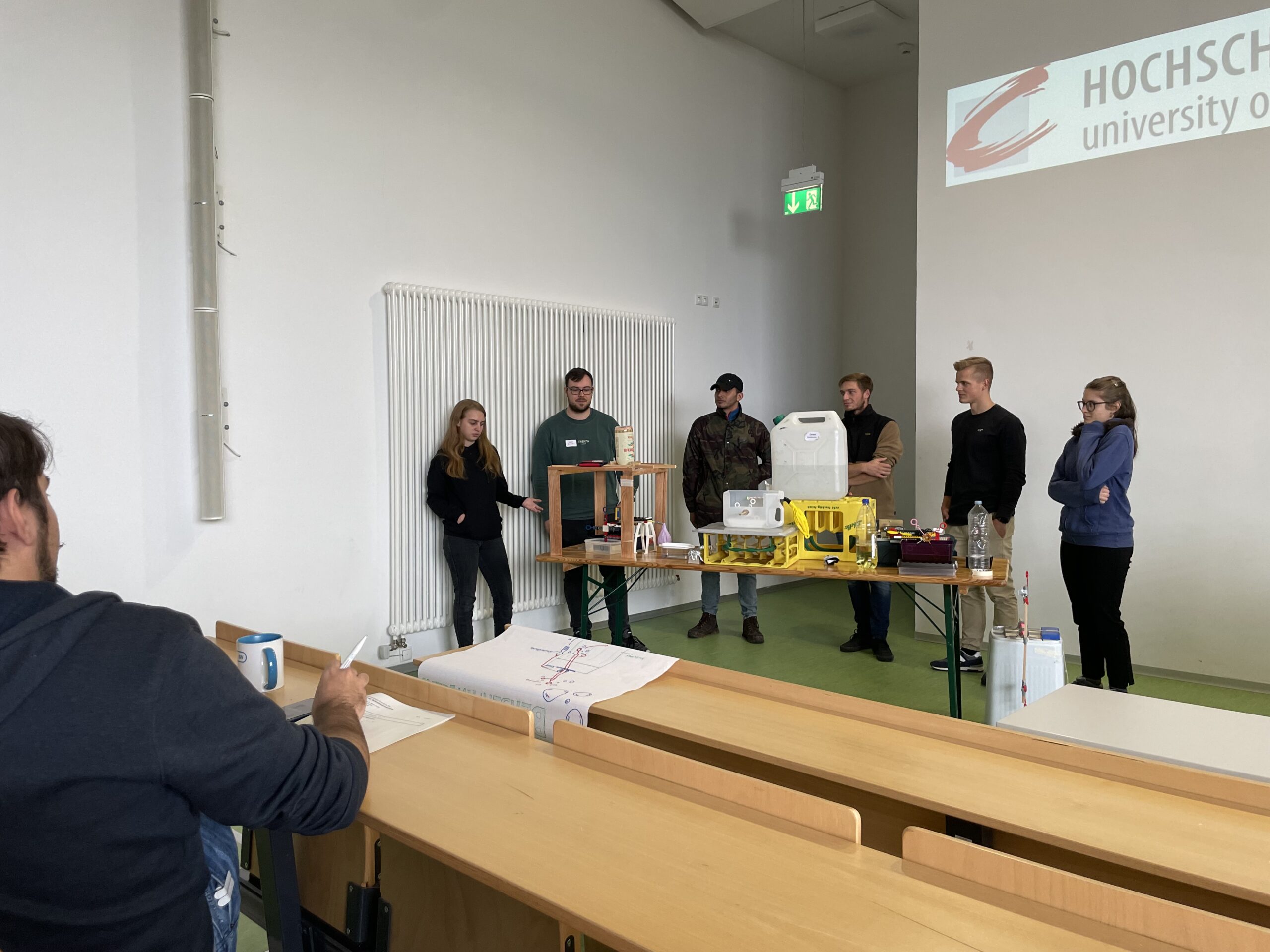 In einem Hörsaal der Hochschule Coburg präsentiert eine Gruppe von Personen auf einem langen Tisch verschiedene Geräte. Eine Person schreibt auf ein großes Blatt Papier, während der Projektor im Raum das Logo der Hochschule an die Wand projiziert.