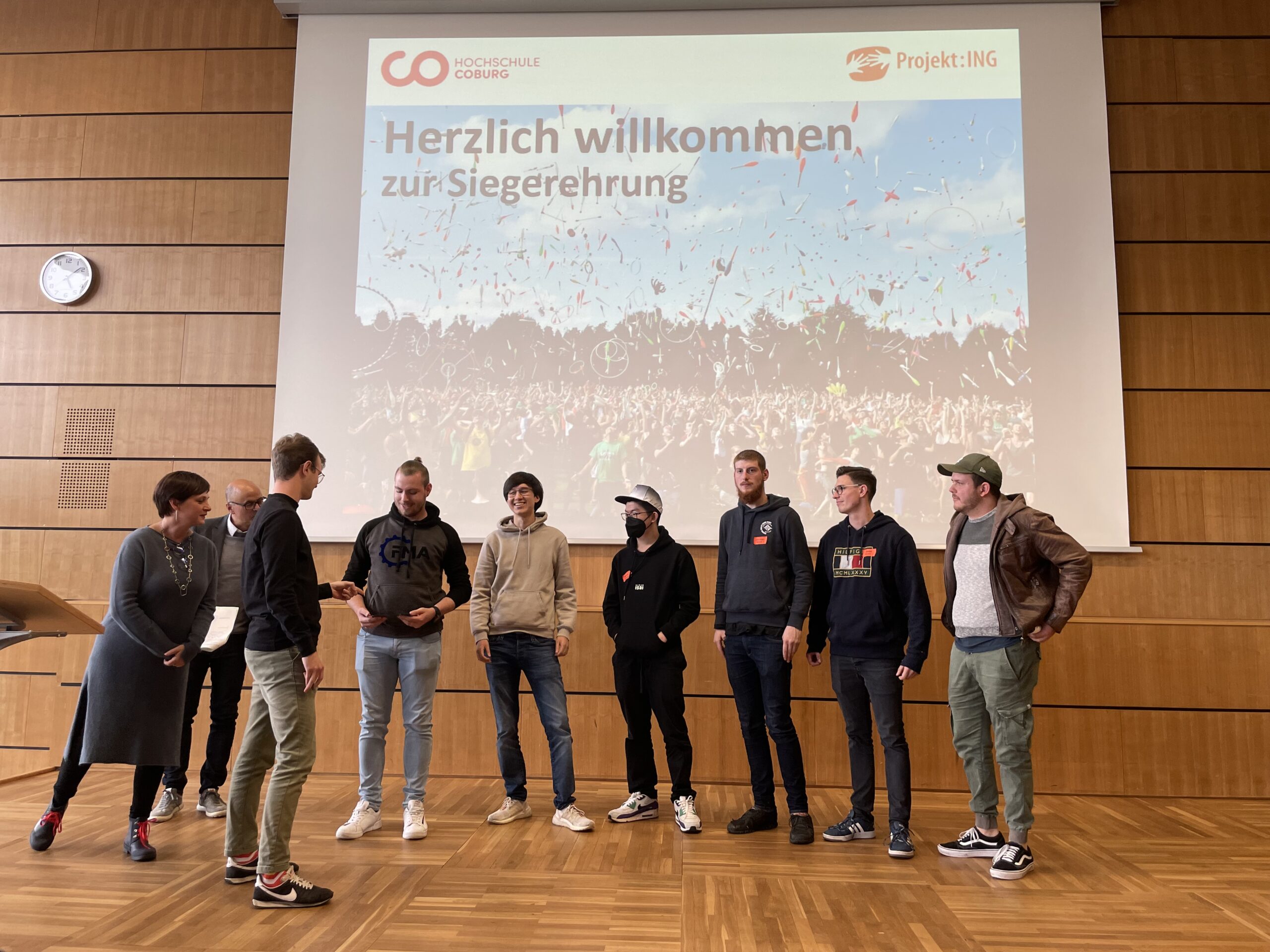 Eine Gruppe von Menschen steht auf einer Bühne vor einer großen Leinwand mit dem Text „Herzlich willkommen zur Siegerehrung“ und den Logos der Hochschule Coburg. Der Raum ist mit eleganten Holzvertäfelungen versehen und einige Personen sitzen dort. Eine Person hält mit einem Mikrofon in der Hand eine Ansprache an die Versammlung.