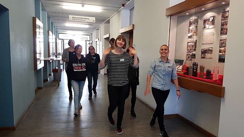 Eine Gruppe von fünf Personen geht einen Flur der Hochschule Coburg entlang. Ihre Gesichter strahlen, als sie an den Vitrinen an den Wänden vorbeigehen. Eine Person winkt fröhlich in die Kamera.