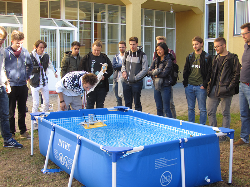 Eine Gruppe von Menschen hat sich im Freien um ein blaues aufblasbares Schwimmbecken an der Hochschule Coburg versammelt. Eine Person beugt sich vor und demonstriert oder justiert ein kleines Objekt, das im Wasser schwimmt. Es scheint sich um eine zwanglose oder pädagogische Veranstaltung zu handeln.