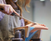 In einer Werkstatt legt eine Person eine Schürze an und formt vorsichtig mit einer Raspel ein Stück Schuhleisten auf der Werkbank. Die handwerkliche Arbeit erinnert an die sorgfältigen Techniken, die an der Hochschule Coburg gelehrt werden.