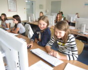 Eine Gruppe junger Studierender sitzt in einem Hörsaal der Hochschule Coburg an Tischen mit Computern. Zwei Mädchen im Vordergrund lächeln und schauen auf einen Monitor. Der Raum, der mit mehreren Computerstationen ausgestattet ist, strahlt eine helle, einladende Atmosphäre aus, die perfekt zum Lernen ist.