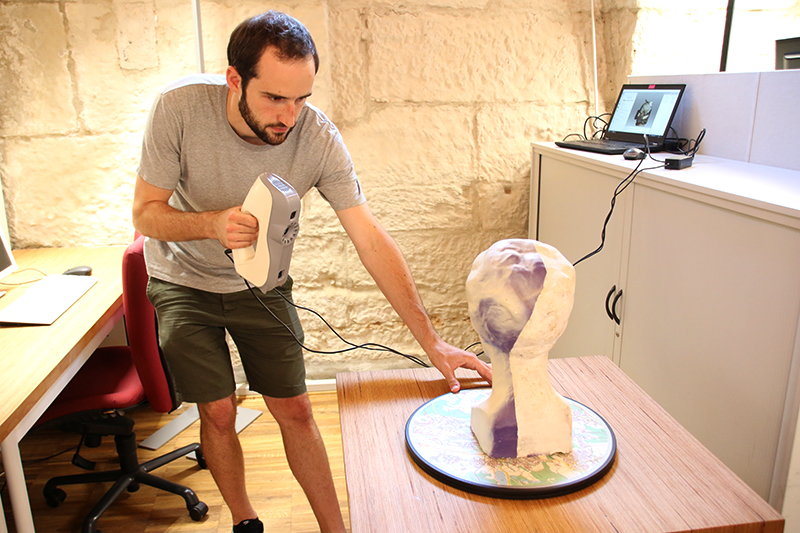 Eine Person erfasst mit einem 3D-Scanner die Details einer Büste auf einer rotierenden Plattform. Der Raum, der an die Innovationslabore der Hochschule Coburg erinnert, ist mit einem Schreibtisch, Stühlen und einem Computer ausgestattet, auf dem entsprechende Software angezeigt wird, wodurch eine inspirierende Arbeitsatmosphäre entsteht.
