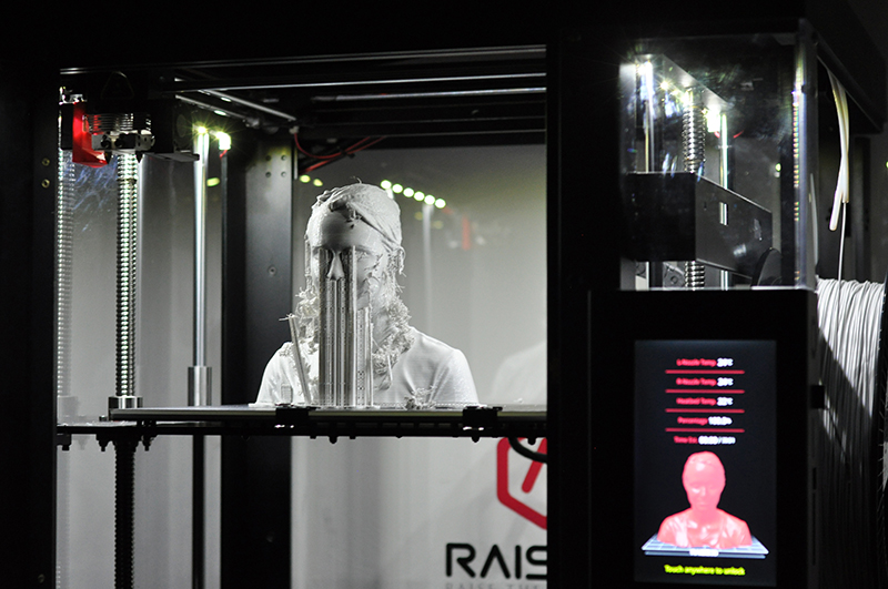 Ein 3D-Drucker an der Hochschule Coburg fertigt eine Büste einer Person. Die teilweise fertige Figur ist im Inneren der Maschine sichtbar, während eine digitale Anzeige den Fortschritt und Druckdetails anzeigt.