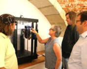 Vier Personen versammeln sich um einen 3D-Drucker an der Hochschule Coburg. Eine Frau deutet auf die Maschine, während zwei Männer mit langen Haaren und ein weiterer Mann mit Brille in einem Raum mit gewölbter Ziegeldecke aufmerksam zuschauen.