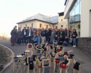 Auf einem Weg neben einem modernen Gebäude der Hochschule Coburg posiert eine große Gruppe von Menschen für ein Foto. Davor sind unter freiem Himmel mehrere kunstvolle Holzskulpturen mit unterschiedlichen Aufsätzen auf dem Boden arrangiert.