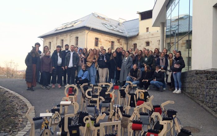 Auf einem Weg neben einem modernen Gebäude der Hochschule Coburg posiert eine große Gruppe von Menschen für ein Foto. Davor sind unter freiem Himmel mehrere kunstvolle Holzskulpturen mit unterschiedlichen Aufsätzen auf dem Boden arrangiert.