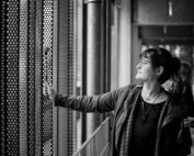 Eine Frau mit Brille und Kurzhaarfrisur steht in einem Gebäude der Hochschule Coburg und berührt sanft ein perforiertes Metallgitter an einer Gebäudewand. Sie wirkt nachdenklich und blickt durch das Muster im Gitter hinaus. Die Szene ist in Schwarzweiß festgehalten.