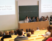 In einem Hörsaal der Hochschule Coburg hält ein Dozent eine Rede an einem Podium vor einer großen Tafel. Die Studenten sitzen in Reihen und hören aufmerksam zu. Auf zwei Projektionsflächen ist ein Text auf Deutsch zu sehen: „Lebensraum Schule – vom Flur zum Foyer“.