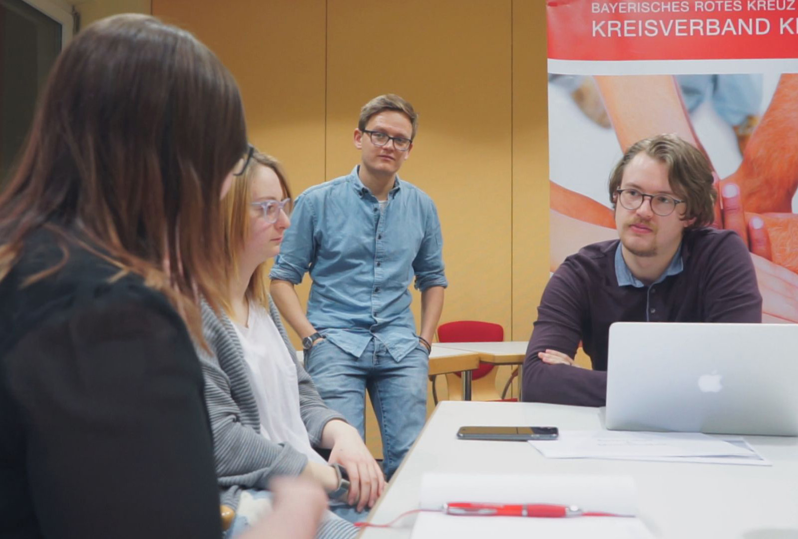 Vier Personen diskutieren in einem Büro. Drei sitzen mit Laptop und Dokumenten an einem Tisch, eine Person steht im Hintergrund. Ein Banner des Bayerischen Roten Kreuzes und der Hochschule Coburg ist zu sehen und spiegelt ihre Zusammenarbeit wider.