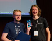Zwei Menschen lächeln auf der Bühne der Hochschule Coburg. Einer hält ein Zertifikat in der Hand und zeigt stolz ein Telefon, auf dem das Gesicht einer Frau während eines Videoanrufs zu sehen ist. Beide tragen Lanyards und stehen vor dem Hintergrund eines beleuchteten Präsentationsbildschirms.